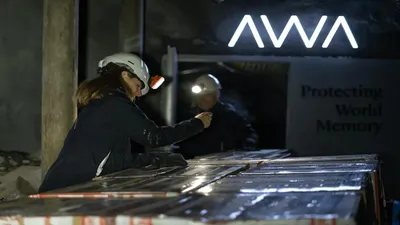 A person wearing a white hard hat with a headlamp and a dark jacket is bent over a long table, meticulously arranging or inspecting numerous rectangular, flat items. Their hands are gloved. In the background, another person with a headlamp is visible in what appears to be a dark, cavernous space. Above them, a glowing white sign reads "AWA" and below it, text says "Protecting World Memory." The overall setting suggests an underground storage facility or archive.
